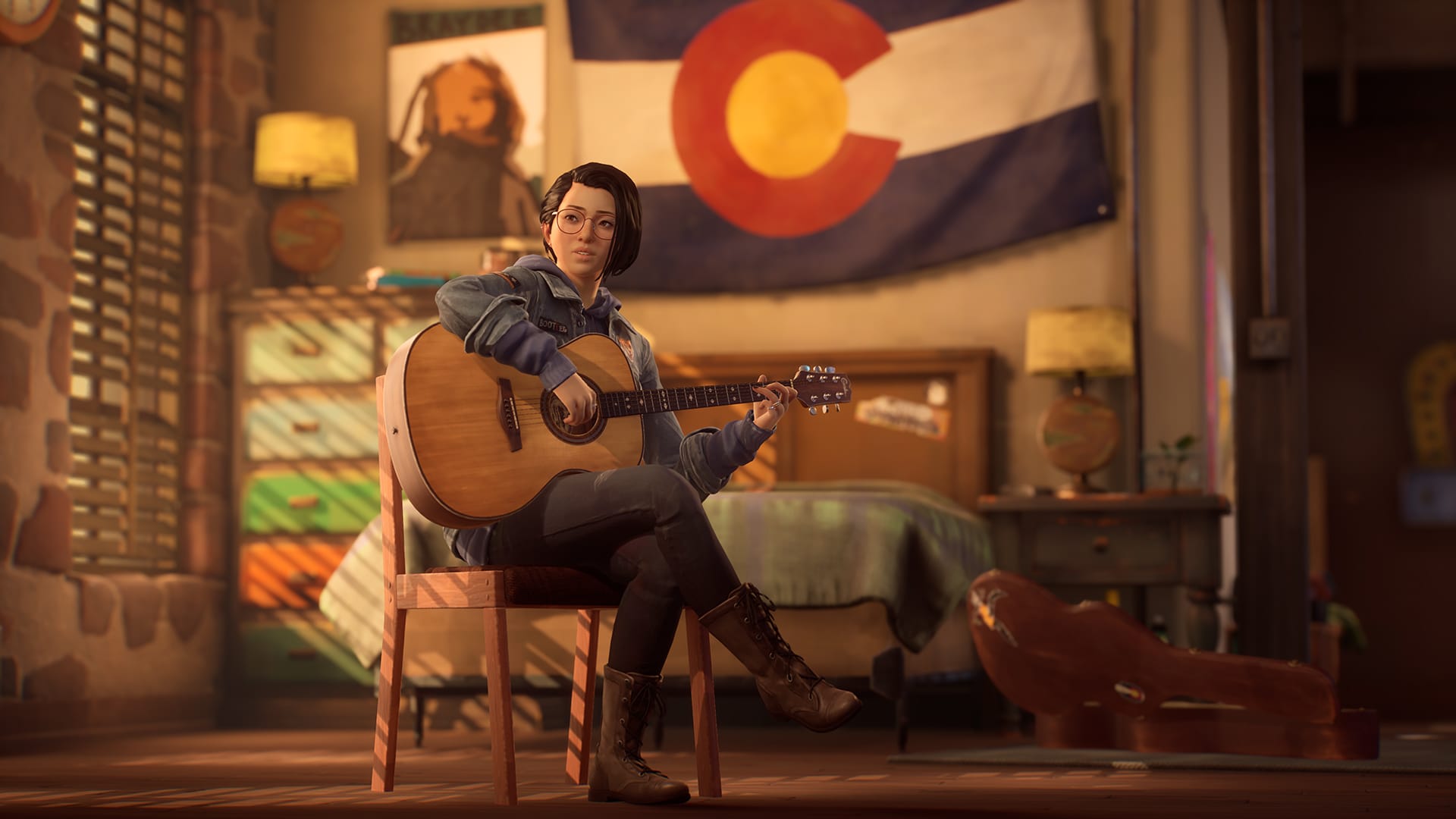 Alex Chen sits crossed legged on a chair, strumming a guitar. She sits in front of a Colorodo State flag and an empty bed.